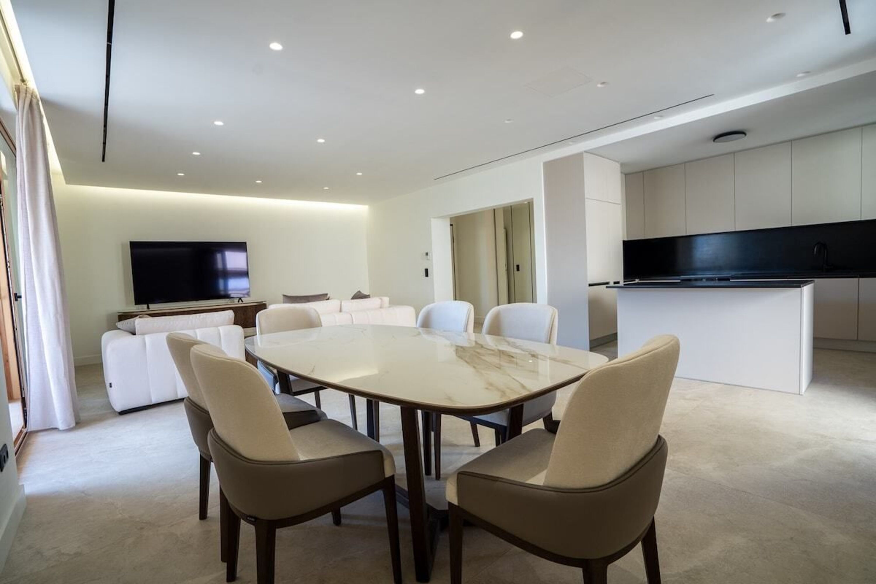 A modern living room with a large TV, a dining table with chairs, a sofa, and a kitchen area with cabinets.