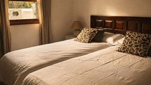Quarto com duas camas, janela com cortinas, mesa de cabeceira com abajur e cabeceira de madeira.