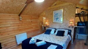A cozy attic bedroom with a wooden sloped ceiling, a bed with a dark headboard, a small desk with a chair, a radiator, and a painting on the wall.