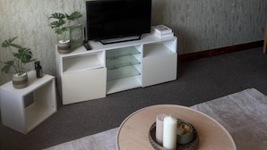 Ett vardagsrum med en platt-TV placerad på en vit TV-bänk, en krukväxt och ett ljus på ett runt bord.