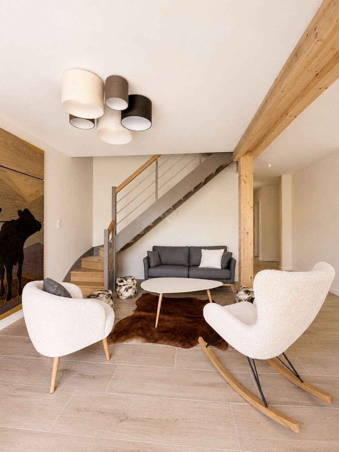 Un salon moderne avec un escalier, un canapé gris, deux fauteuils blancs et un tapis marron.