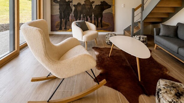 Una sala de estar moderna con un ventanal grande, techo de madera, una escalera y un cuadro de vacas.