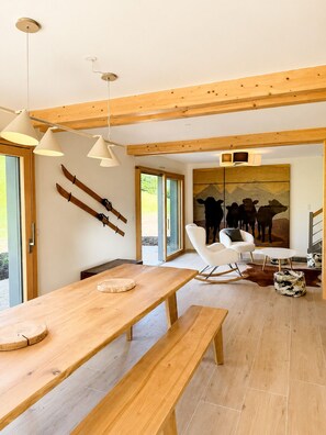 Un intérieur moderne avec des poutres en bois, une table à manger avec des bancs, une chaise blanche et un tableau représentant des vaches.