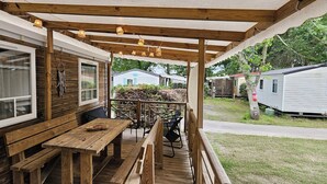 Une terrasse en bois avec une table et des bancs, un espace couvert agrémenté de guirlandes lumineuses, et une vue sur les maisons mobiles voisines.