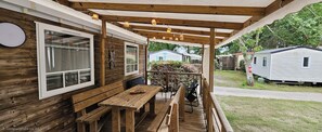 Eine Holzterrasse mit Tisch und Bänken, ein überdachter Bereich mit hängenden Lichtern und Blick auf angrenzende Mobilheime.