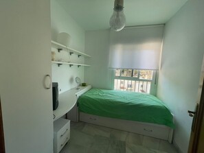 Une petite chambre à coucher, bien rangée, avec un lit simple, un bureau et une fenêtre donnant sur l’extérieur.
