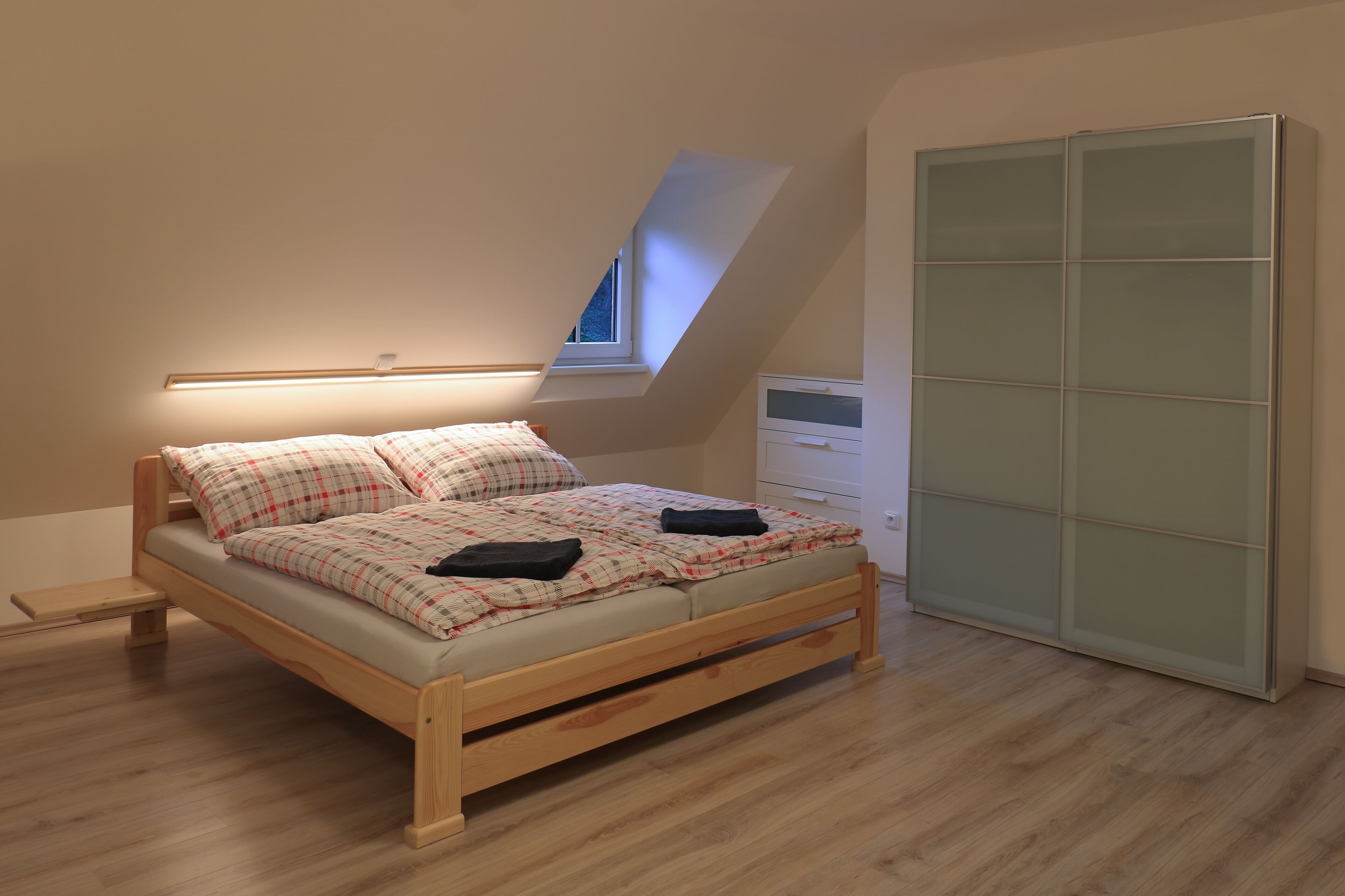Un dormitorio con techo inclinado, una cama, una mesita de noche, un armario y una ventana.
