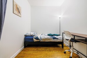 Ett litet rum med en enkel säng, ett skrivbord och en stol, en golvlampa och en inramad tavla på väggen.