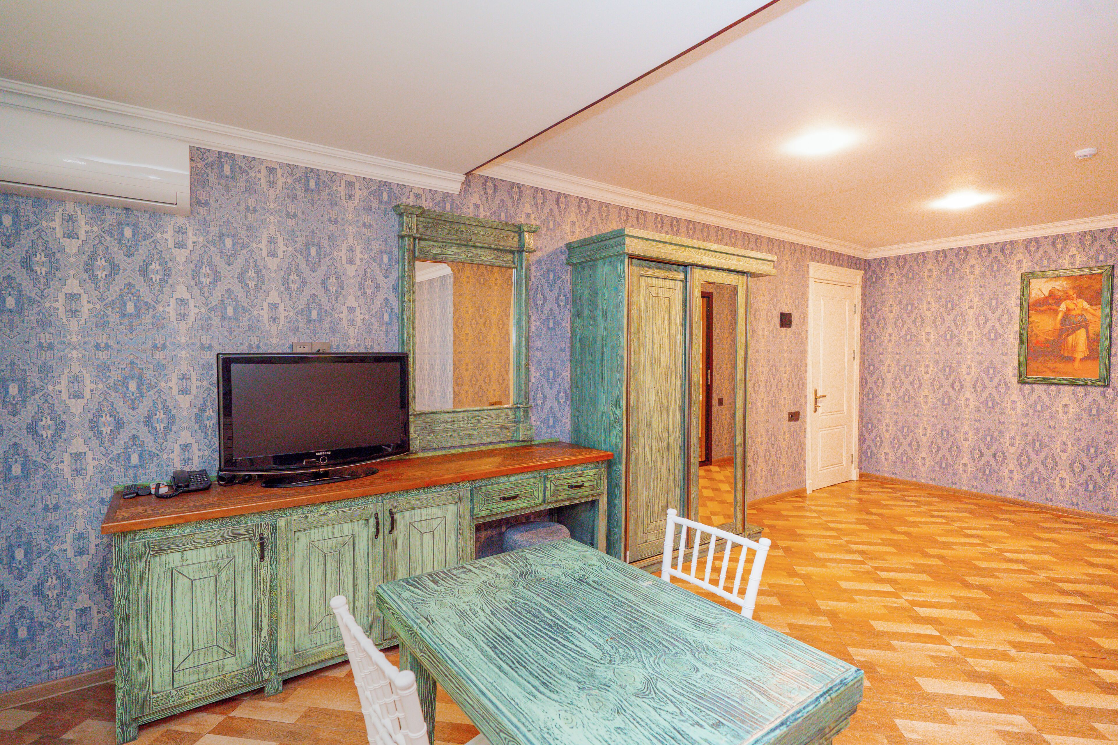 Una stanza con carta da parati decorata, una televisione su un mobile in legno, una sedia bianca e una porta che dà su un'altra stanza.
