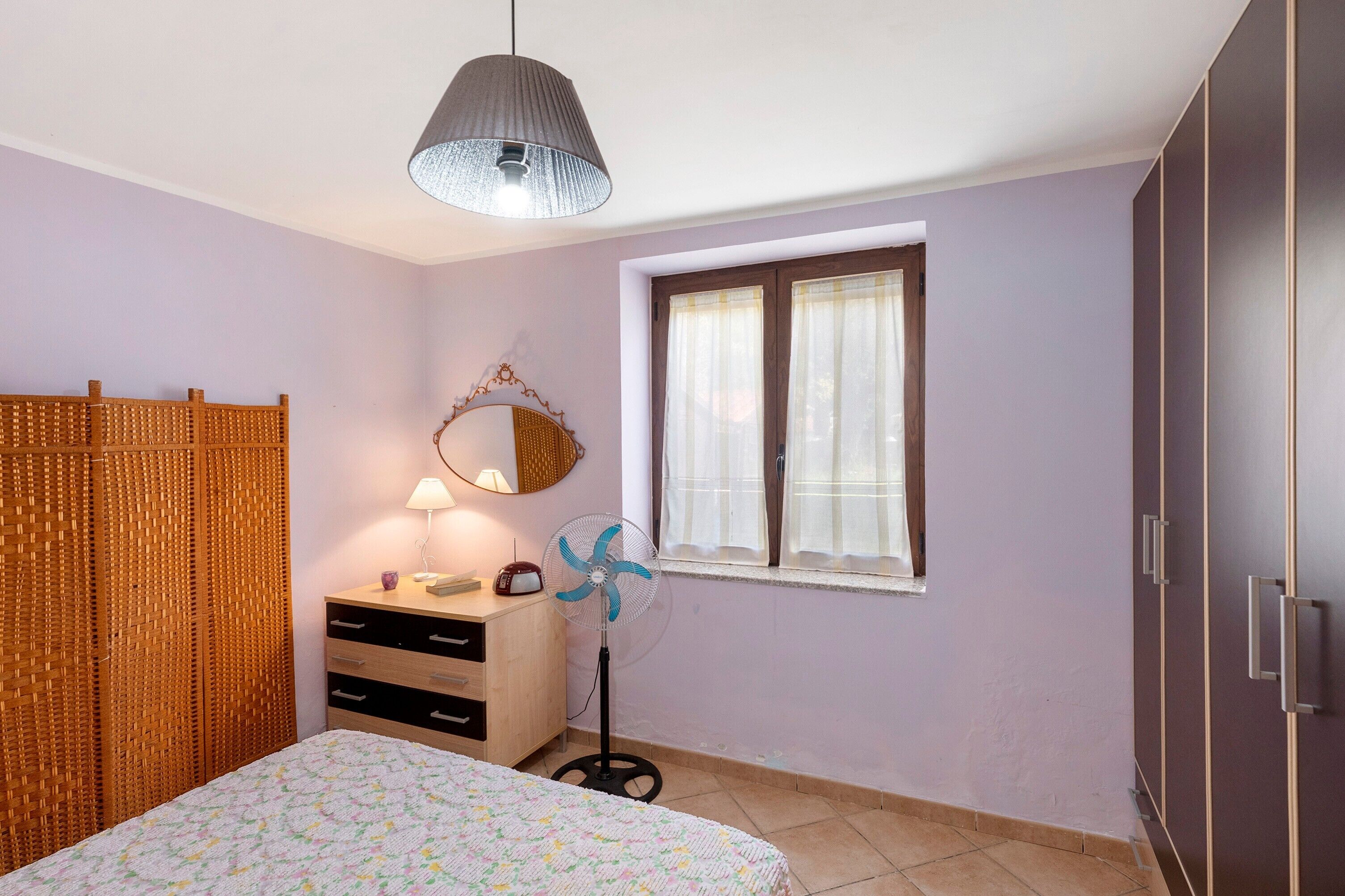 Et soveværelse med seng, et natbord med lampe, en ventilator, et vindue med gardiner og et klædeskab.