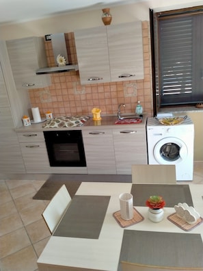 A modern kitchen with a built-in oven, a washing machine, and a dining area with a table set for two. - La Grande Quercia, relaxation in the heart of Cilento (Perdifumo)