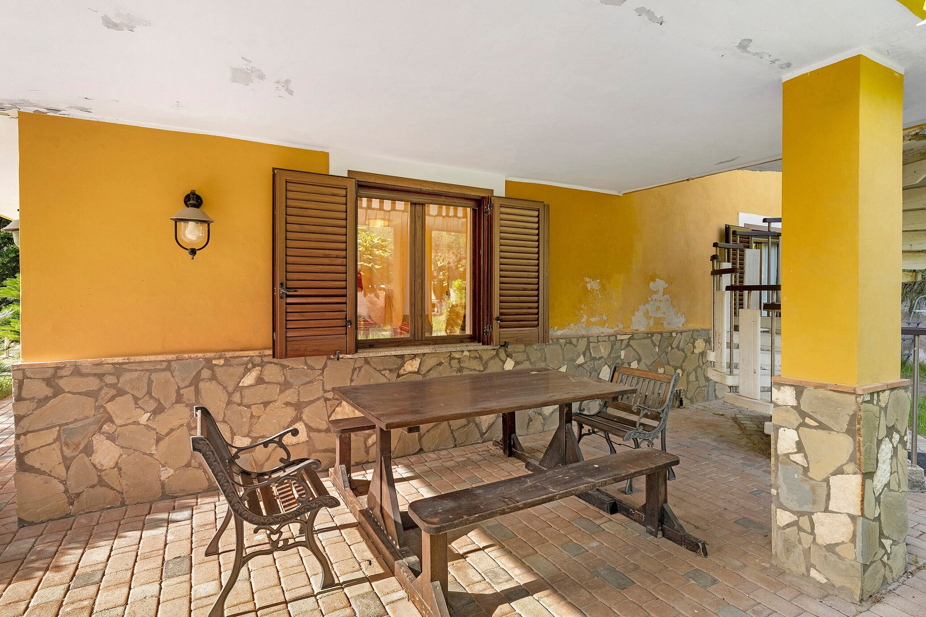 Une terrasse extérieure avec un mur en pierre, une table en bois, des bancs et une fenêtre avec des volets.