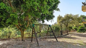 Un'area esterna pavimentata con altalene, circondata da alberi e una recinzione.
