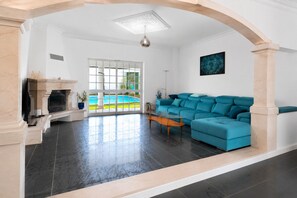 A spacious living room with a blue sofa, a fireplace, and a view of a swimming pool. - Villa Magali – Swimming pool, golf and beach (Charneca de Caparica)