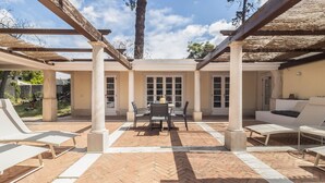 Un patio al aire libre con suelo de ladrillo, sillones blancos, una mesa con sillas y una zona cubierta con columnas.