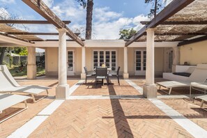 Eine Außenpatio mit rotem Backsteinboden, weißen Lounge-Sesseln, einem Tisch mit Stühlen und einem überdachten Bereich mit Säulen.