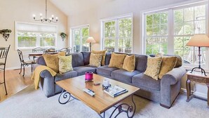 A living room with a sectional sofa, a coffee table, a dining area with a table and chairs, and large windows with a view of greenery. - Perfect Pocono Retreat / Golf / FirePit /Swimming Pool/BBQ near Waterparks (Tobyhanna)