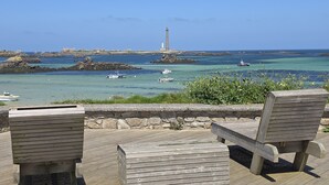 Eine Holzterrasse mit zwei Stühlen und einer Bank mit Blick auf eine Küstenlandschaft, wo Boote und ein Leuchtturm zu sehen sind.