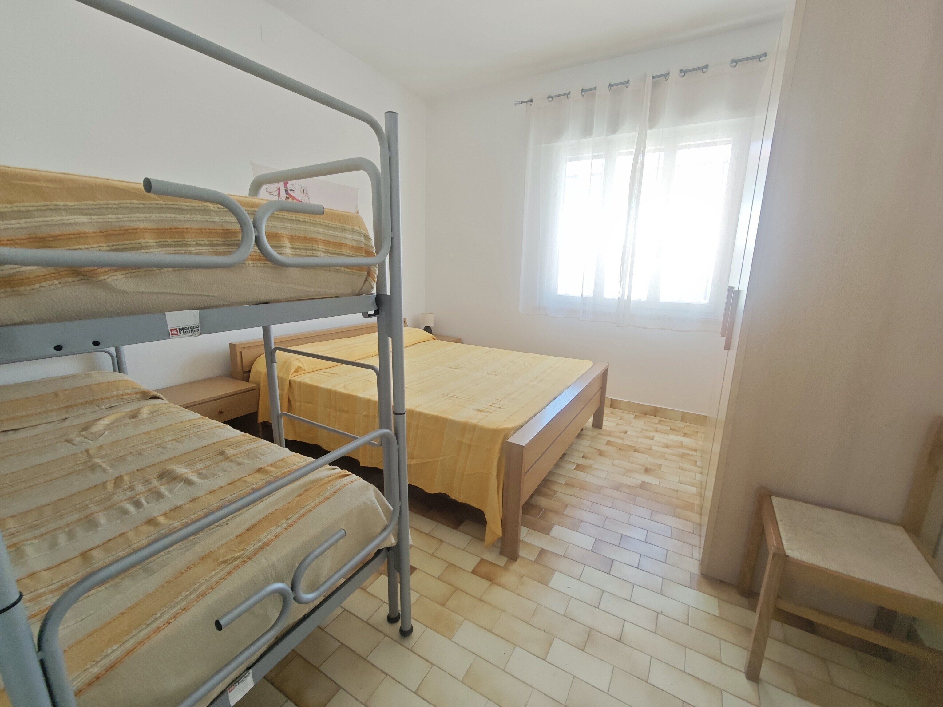 Ein Schlafraum mit Etagenbetten, einem Einzelbett, einem Holzstuhl und einem Fenster mit Vorhängen.