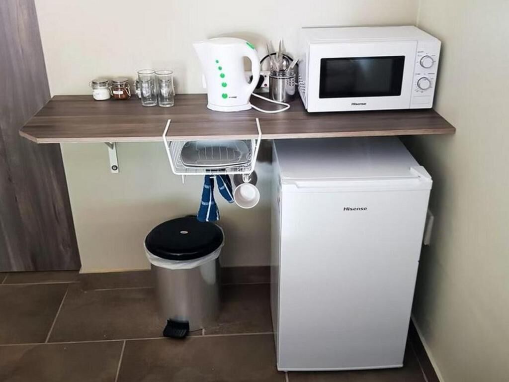 Una kitchenette compacta con microondas, un refrigerador pequeño, un cesto de basura y una alacena con utensilios de cocina.