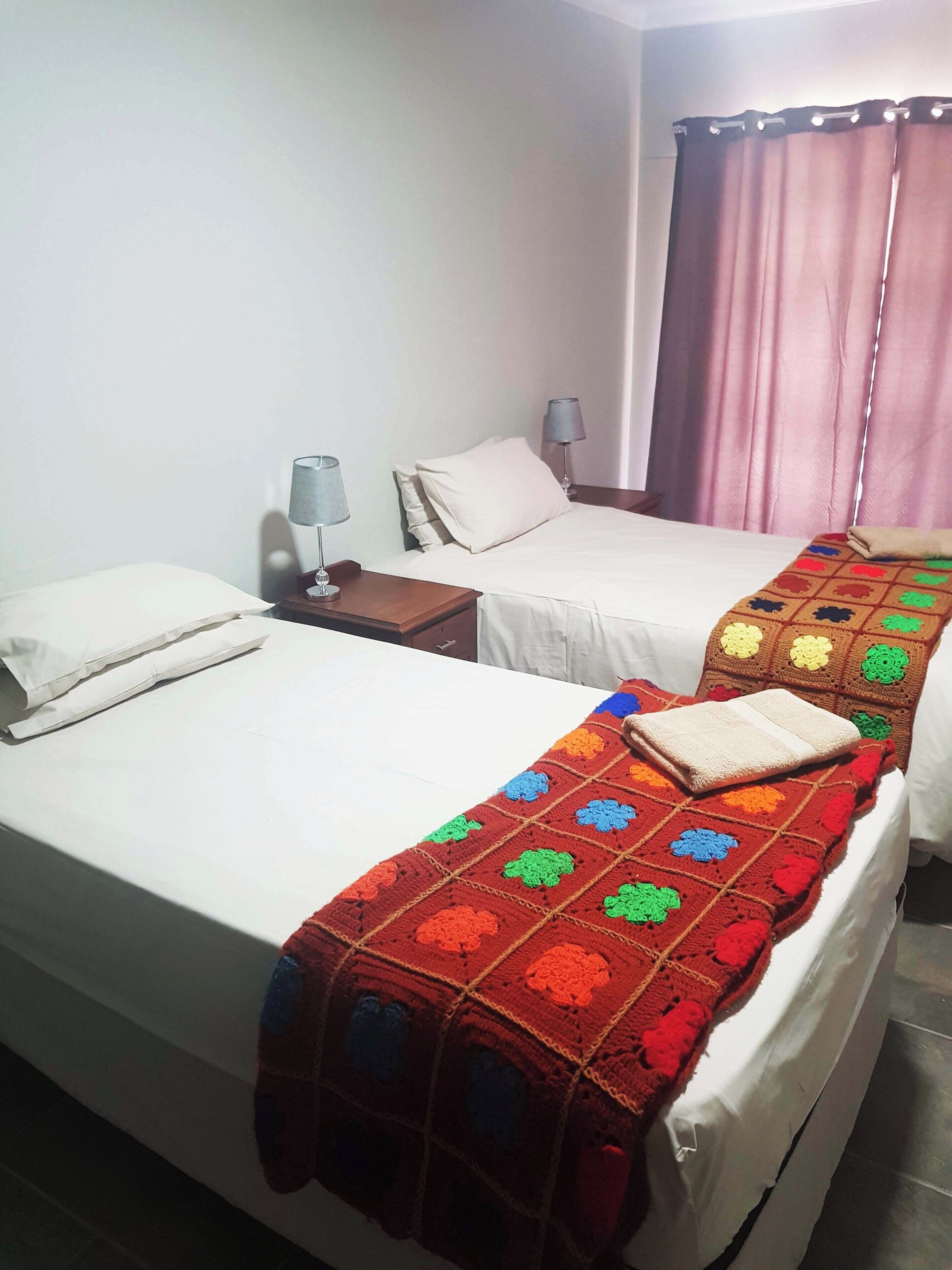 Habitación de hotel con dos camas individuales, una mesita de noche con lámpara, una ventana con cortinas y una manta colorida sobre una de las camas.