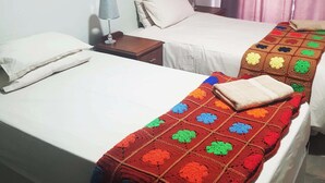 Habitación de hotel con dos camas individuales, una mesita de noche con lámpara, una ventana con cortinas y una manta colorida sobre una de las camas.