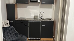 Angolo cottura compatto con forno a microonde, lavello e pensili neri. Nella stanza, un letto e una sedia.
