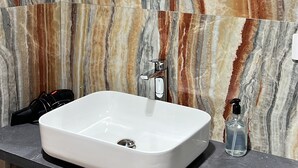 Un bagno con parete in marmo, uno specchio rotondo, un lavabo bianco, un dispenser per sapone e un cestino.