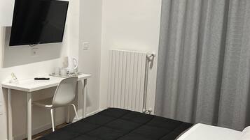 A hotel room with a bed, a desk with a chair, a television, a radiator, and a window with curtains.