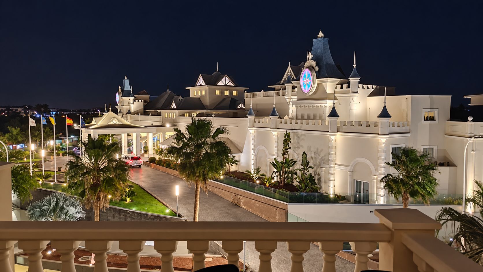 Un hotel con un edificio central grande, varias estructuras más pequeñas y un área de entrada bien iluminada con palmeras.
