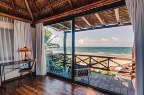 Une piĂšce dotĂ©e dâun plancher en bois, dâune table avec une lampe, dâune chaise et dâune grande porte vitrĂ©e donnant sur une plage avec une terrasse en bois et des chaises longues.