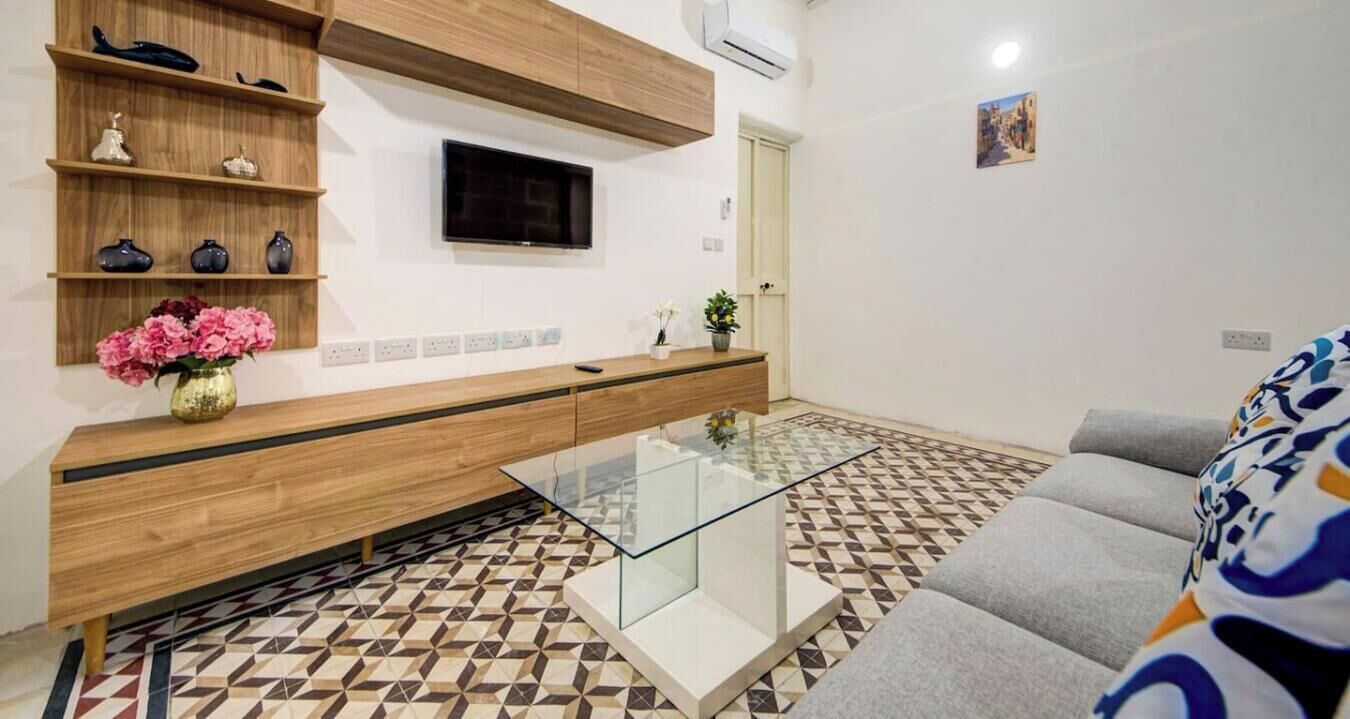 A modern living room with a sofa, a glass coffee table, a TV mounted on the wall, and a patterned floor.