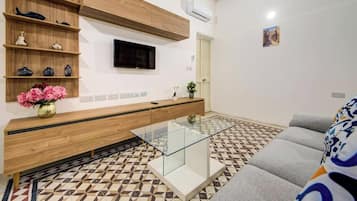 A modern living room with a sofa, a glass coffee table, a TV mounted on the wall, and a patterned floor.