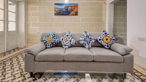 Un canapé gris avec des coussins à motifs, un sol carrelé, un tableau accroché au mur et une porte avec un panneau indiquant la sortie.
