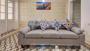 A grey sofa with patterned cushions, a tiled floor, a wall-mounted picture, and a door with an exit sign.