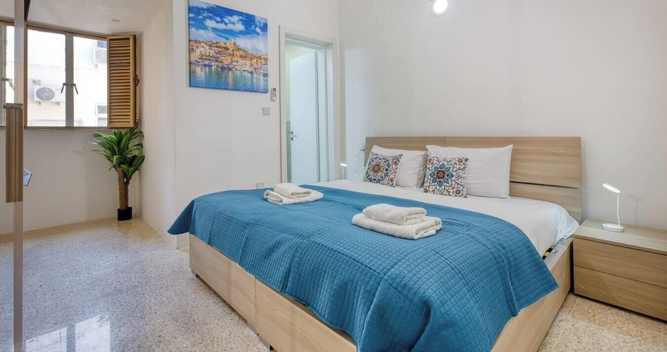 A bedroom with a bed, a nightstand, a potted plant, a painting, and a door.