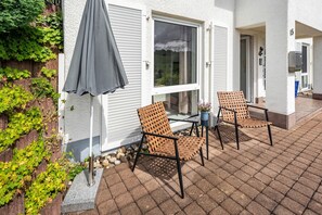 Un patio con due sedie, un tavolo, un ombrellone grigio e un edificio bianco con una finestra e una porta.