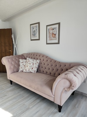 Un canapé capitonné agrémenté de coussins à motifs floraux, dans une pièce dotée d’un parquet en bois clair et de murs blancs.