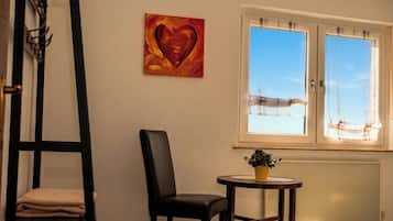 Ein Zimmer mit einem schwarzen Stuhl, einem kleinen Tisch mit einer Pflanze, einem Fenster mit Vorhängen und einem Herzbild an der Wand.