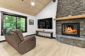 A modern living room with a stone fireplace, a recliner, a large window, and a flat-screen TV. - 2-bedroom house in adorable Ashford with, AC, EV Charger, Hot Tub Wifi (Ashford)