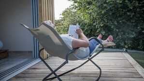 Iemand ligt ontspannen in een stoel met een boek, op een houten terras met een glazen deur en een tuin op de achtergrond.