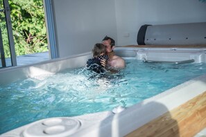 A hot tub with a wooden edge and a metal handle, surrounded by a window with a view of greenery.