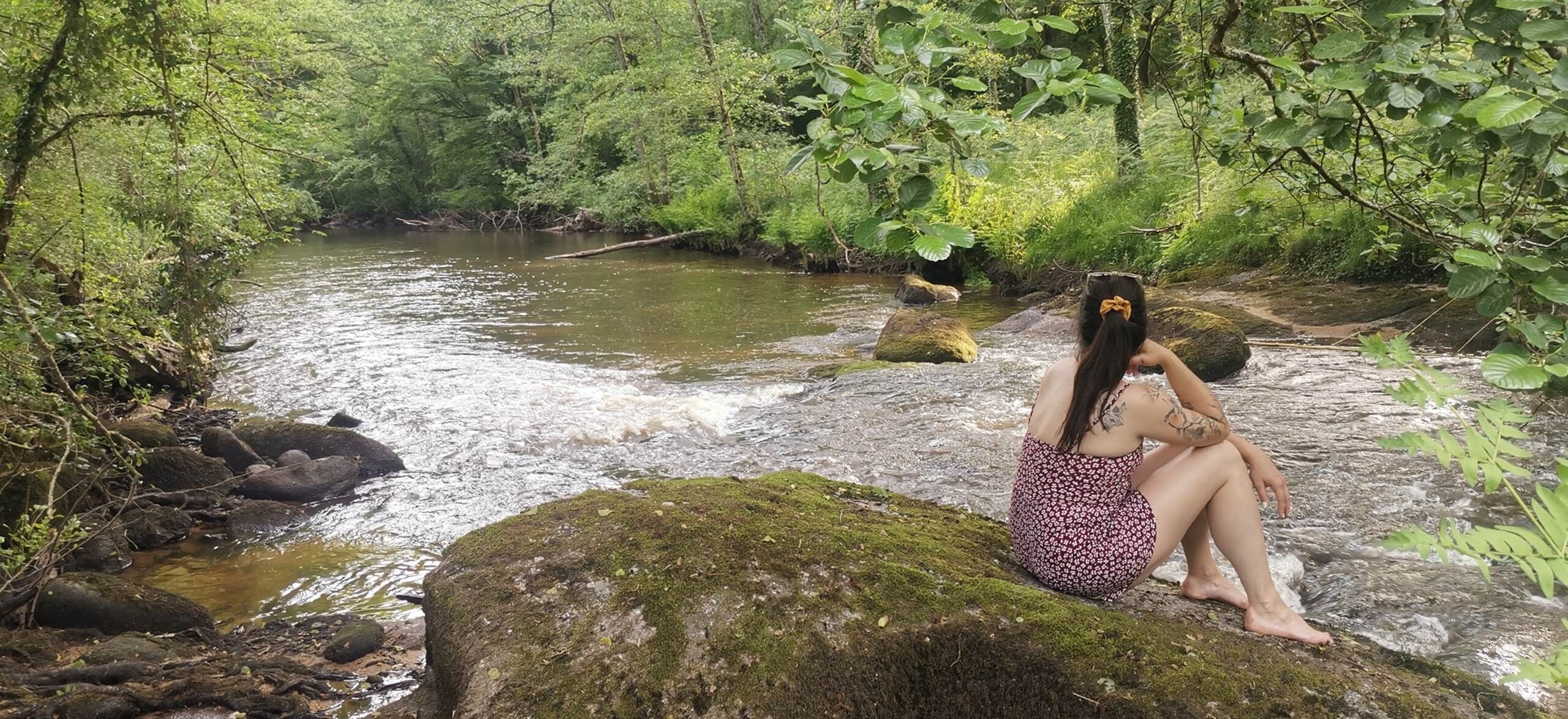 A person sitting on a moss-covered rock by a flowing river in a forested area.