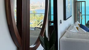 Ein modernes Wohnzimmer mit einem großen Spiegel, einer Topfpflanze und Blick auf das Meer und Gebäude.