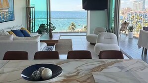 Ein modernes Wohnzimmer mit großem Fenster mit Meerblick, ein Essbereich mit Marmortischen und einem Deckenventilator.