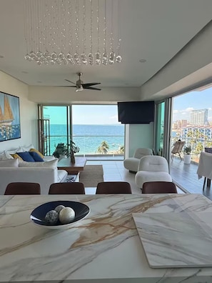 A modern living room with a large window overlooking the ocean, a dining area with marble tables, and a ceiling fan. - Luxury 1-bedroom apartment at Icon Vallarta 2-801 with an ocean view (Puerto Vallarta)