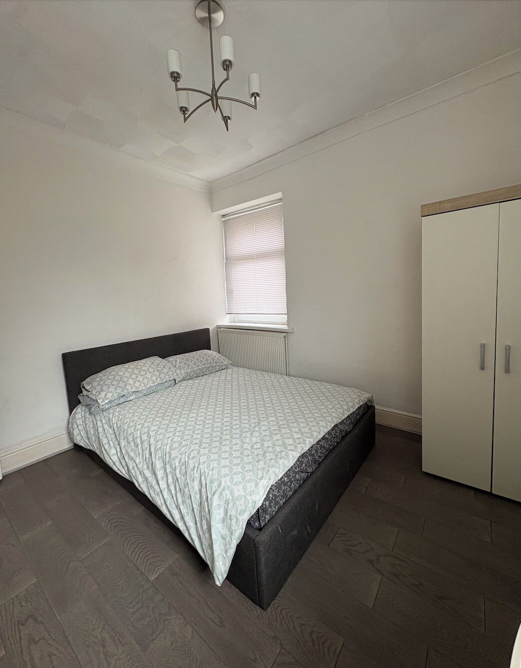 A bedroom with a bed, a wardrobe, a window with blinds, and a chandelier.