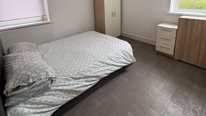 Et soveværelse med seng, garderobeskab, kommode, et vindue med skodder og en loftlampe.