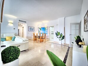 Ein modernes Wohnzimmer mit einem weißen Sofa, einem Esstisch mit Stühlen, einer Topfpflanze und Blick in die Küche durch eine offene Tür.