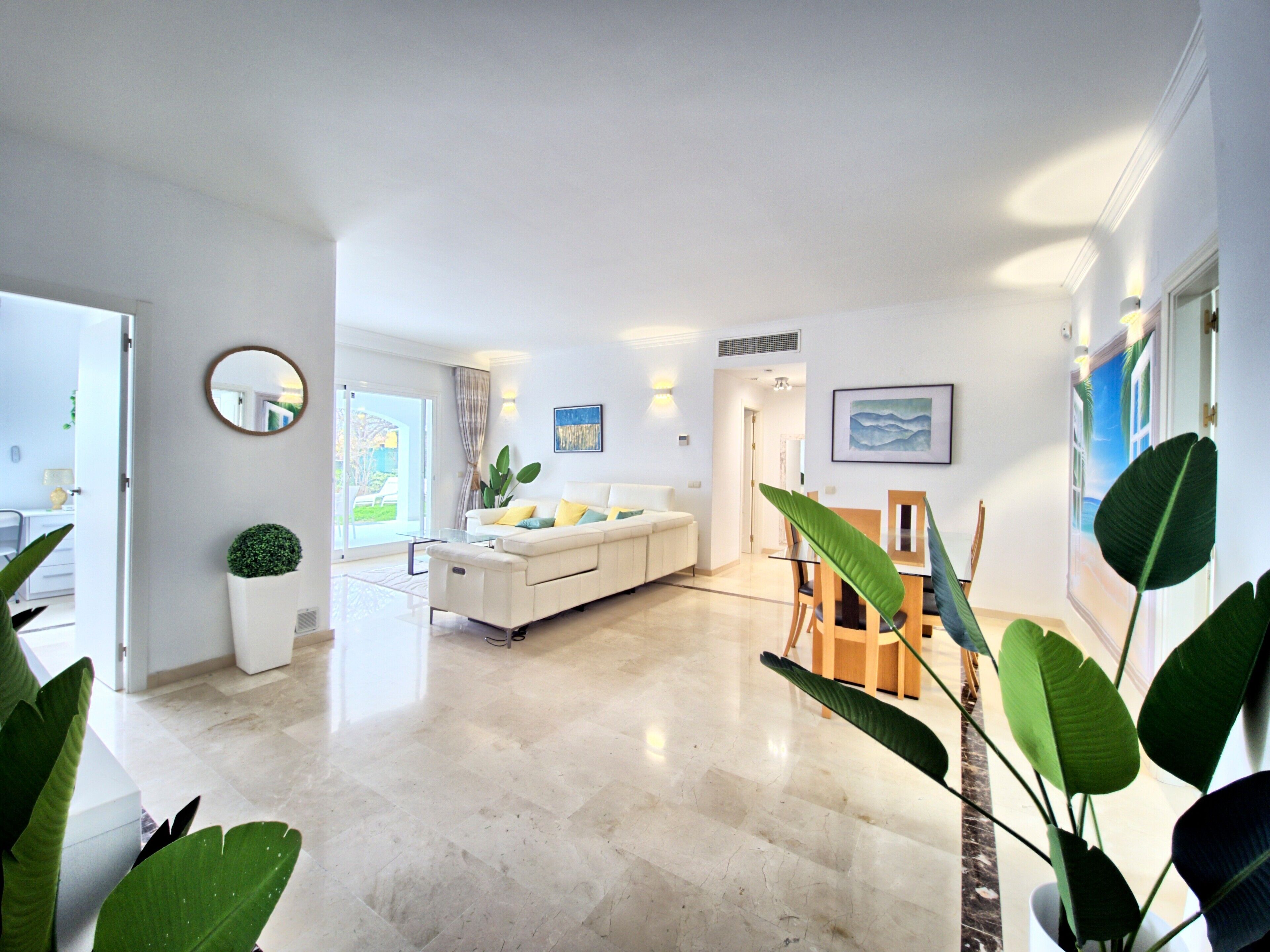 A spacious living area with a sofa, a dining table, and chairs, a large mirror, and a potted plant.